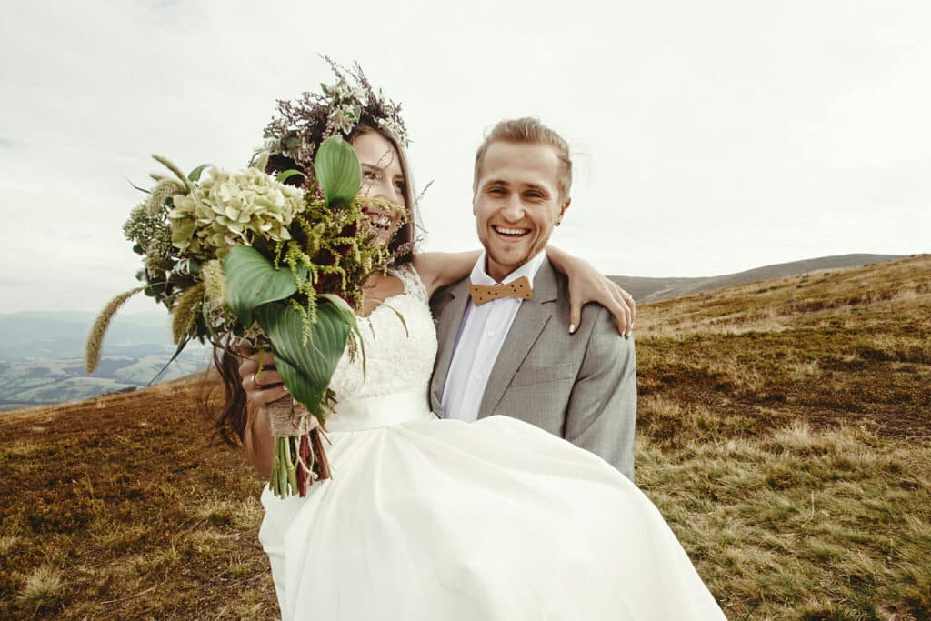 Et smilende brudepar står på en bakke, omgivet af naturens skønhed og glæde på deres store dag.