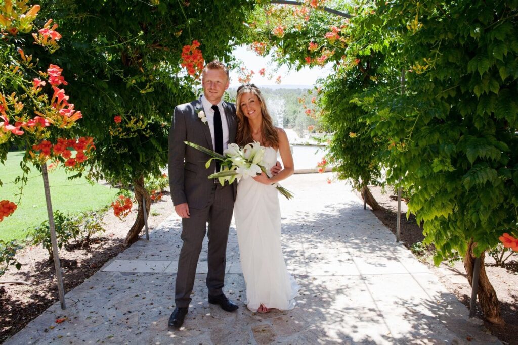 En brud og brudgom står i en blomsterbue, omfavnet af farverige blomster i en romantisk setting i Spanien.